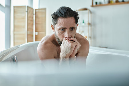 anxious man sitting in bathtub with hands near face during breakdown, mental health awarenessの写真素材