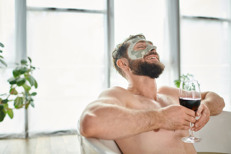 cheerful attractive man with beard and face mask relaxing in bathtub with glass of red wineの写真素材