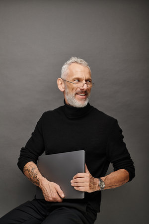 cheerful handsome bearded mature man with glasses sitting on chair with laptop and smiling happily