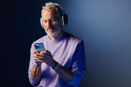 joyful attractive mature man with beard and headphones enjoying music and looking at his phone