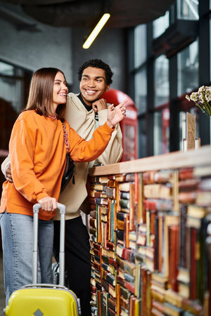 cheerful diverse couple with luggage laughing, woman pointing with finger at hostel reception deskの写真素材
