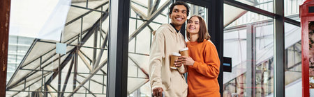 Young diverse couple smiling while holding coffee to go and entering a modern hostel, bannerの写真素材