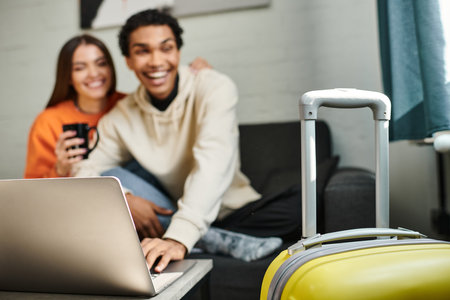 focus on yellow luggage, happy diverse couple traveling together hostel living room and laptop