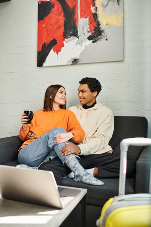 happy interracial couple sitting on a couch in hostel, surrounded by laptop and luggage