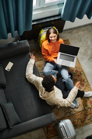 diverse couple lounging on floor, sharing laughter and spending time near luggage in hostel