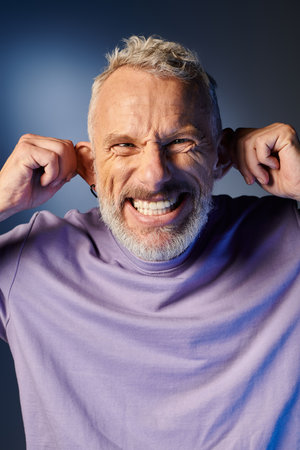 cheerful mature man with gray beard and casual trendy attire grimacing and looking at camera