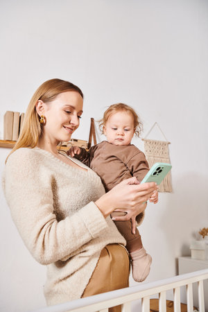young mother with toddler son in hands messaging on mobile phone in nursery room, multitasking woman