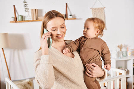 smiling woman with little son in hands talking on mobile phone in nursery room, multitasking mother