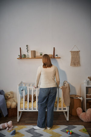 back view of grieving and depressed woman near crib with soft toys un dark nursery room at homeの写真素材