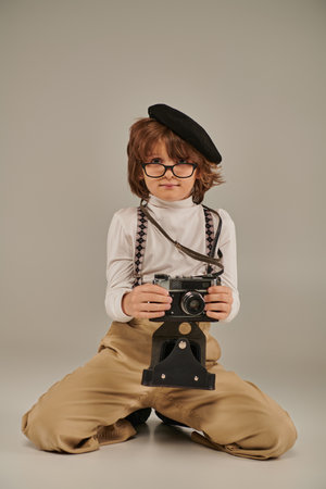 young photographer in beret and glasses holding camera and sitting on floor, boy in suspendersの写真素材