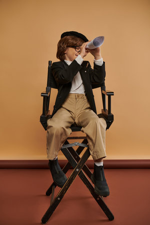 cute boy in stylish clothing sits on director chair and looking through hole in rolled paperの写真素材