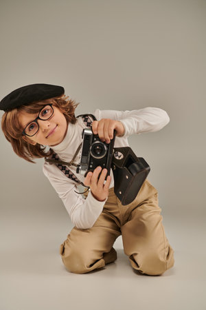 young photographer in beret with retro camera sitting on floor, curious boy in suspendersの写真素材