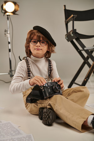 boy in beret holding retro camera and sitting on floor near director chair, young photographerの写真素材