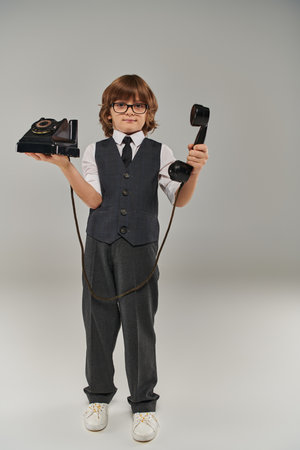 elegant boy in glasses and stylish vest holding retro telephone on grey background, nostalgiaの写真素材