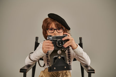 boy in beret and glasses holding vintage camera while sitting on director chair, young photographerの写真素材