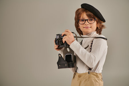 pleased boy, young photographer in beret and suspenders taking photo on retro camera in studioの写真素材