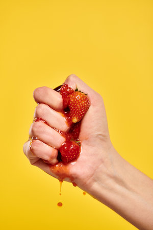 unknown young woman with nail polish squeezing red juicy fresh strawberries on yellow backgroundの写真素材