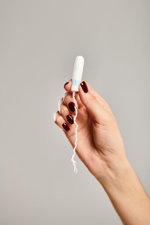 unknown young female model with nail polish holding in hand hygienic tampon on gray background