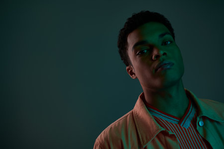handsome african american man in shirt looking at camera in dark studio with green lightの写真素材