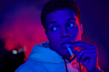 cool african american man holding rolled cigarette near mouth on dark blue background with red light