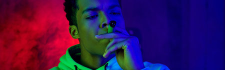 banner of african american young man in hoodie smoking cigar on dark blue background with red light