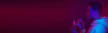 young african american man in hoodie looking at glass bong on dark purple background, banner