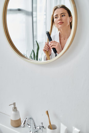 attractive blonde woman in cozy bathrobe using flat iron on her hair and looking at mirrorの写真素材