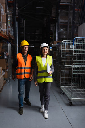 two colleagues with hard hats walking in a well-lit warehouse, supervisor and employeeの写真素材