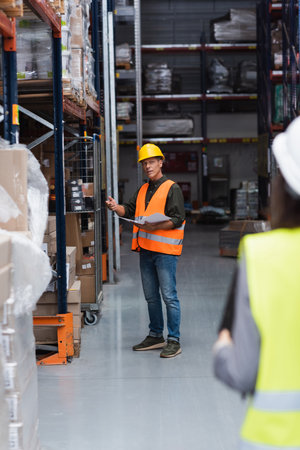 middle aged supervisor in hard hat with paperwork instructing his female employee in warehouse