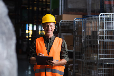 happy middle aged supervisor in hard hat reviewing paperwork in warehouse, logistics and cargoの写真素材