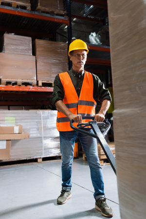 serious middle aged warehouse worker in hard hat transporting pallet with a hand truck