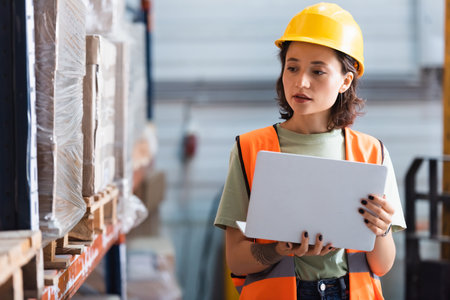 female worker in hard hat and safety vest using laptop while checking inventory in warehouse