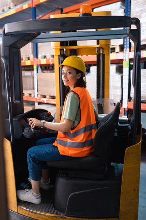 happy female forklift operator in hard hat and safety vest looking at camera in warehouse, logisticsの写真素材