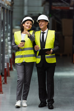 happy logistics workers with hard hats walking with coffee near inventory while inspecting warehouseの写真素材