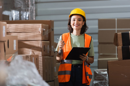 cheerful female warehouse worker in safety vest and hard hat holding clipboard and pen near cargoの写真素材