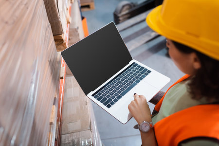 female warehouse worker in hard hat and safety vest using laptop while checking cargo, overheadの写真素材