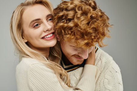 happy and fashionable blonde woman embracing young redhead man on grey, trendy couple relationshipの写真素材