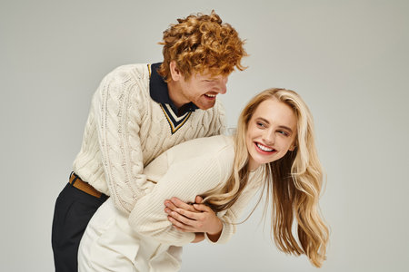 cheerful redhead man hugging excited blonde woman and having fun on grey backdrop, classic fashionの写真素材