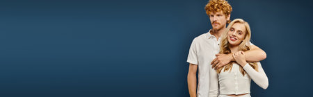 young redhead man embracing joyful blonde woman in white stylish attire on blue backdrop, bannerの写真素材