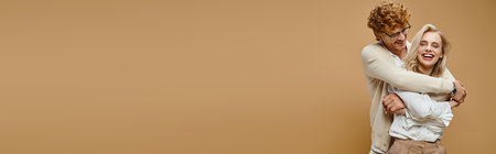 overjoyed redhead man in eyeglasses embracing laughing blonde woman on beige, horizontal banner