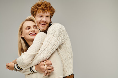 excited redhead man embracing blonde woman in trendy casual clothes on grey, timeless fashionの写真素材