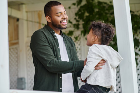 cheerful african american father hugging cute curly son sitting on porch on house in suburbsの写真素材
