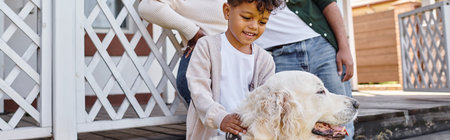 positive african american kid playing with dog near parents on backyard of their house, bannerの写真素材