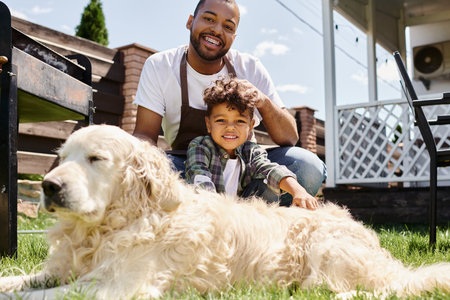 happy african american man in apron touching curly hair of son and sitting near family dog outdoorsの写真素材