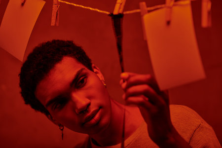 young african american man looking at freshly developed film strip  in a red-lit darkroom, nostalgia