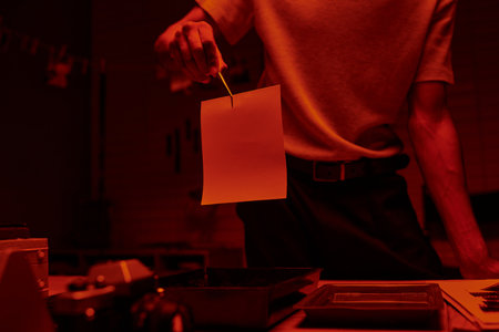 cropped shot of photographer holding tweezers with photo paper in a darkroom with red lightの写真素材