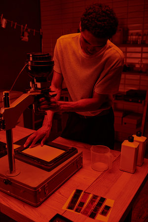 dark skinned photographer focuses on delicate process of enlarging film in darkroom with red lightの写真素材