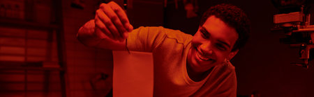 happy african american photographer holding tweezers with developed photo paper in darkroom, bannerの写真素材