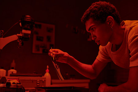 african american photographer holding developed photo paper with tweezers in red toned darkroom