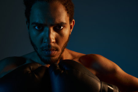 appealing african american man with boxing gloves posing on dark backdrop and looking at camera